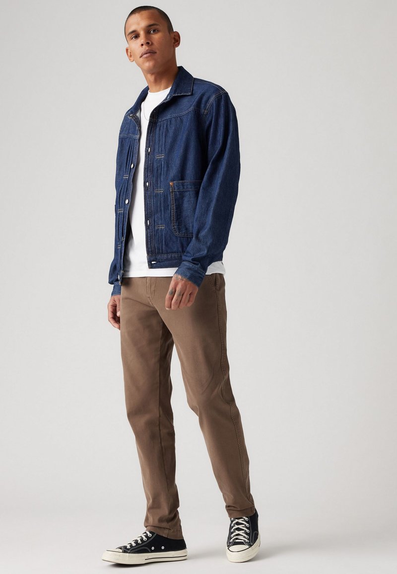 Veste en denim bleu foncé avec fermeture à boutons et poche poitrine, associée à un pantalon marron et des sneakers noires avec semelles et lacets blancs.