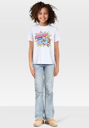 Hvid bomulds t-shirt med grafik af "The Powerpuff Girls" i livlige farver, parret med lyseblå flagrende jeans og beige sko.