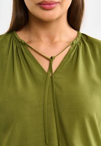 Žalia bluzonė su susiraukšlėtu kaklo apdailos detalėm, minkšta medžiaga, V formos iškirpė ir rišimo uždarymas priekyje. Lygi tekstūra ir paprastas dizainas.