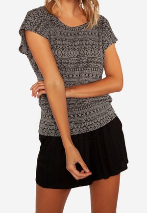 Femme portant un haut gris à manches courtes à motifs et un short noir, posant avec un bras croisé et l'autre main détendue.
