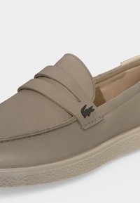 Mocassin beige en cuir à enfiler avec une semelle texturée et un petit logo crocodile en métal foncé sur la bride latérale.