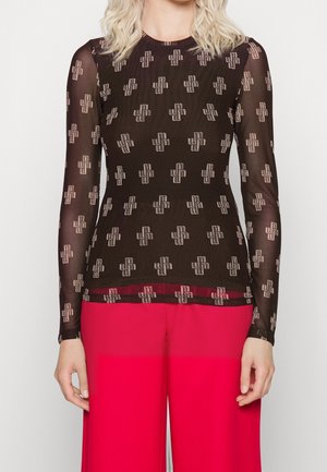 Femme portant un haut noir transparent à manches longues avec des motifs géométriques blancs et un pantalon rouge vif, se tenant devant un fond uni.