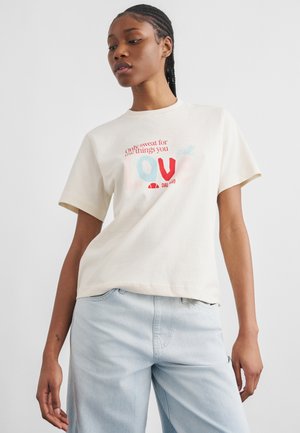 Γυναίκα με πλεγμένα μαλλιά που φοράει ένα ανοιχτό μπεζ T-shirt με το κείμενο «Μόνο ιδρώτας για τα πράγματα που ΑΓΑΠΑΣ» και ανοιχτό μπλε τζιν, μπροστά από μια απλή φόντο.