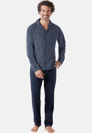 Man standing barefoot wearing navy blue patterned long-sleeve shirt and solid navy blue pants, smiling with hand on hip.