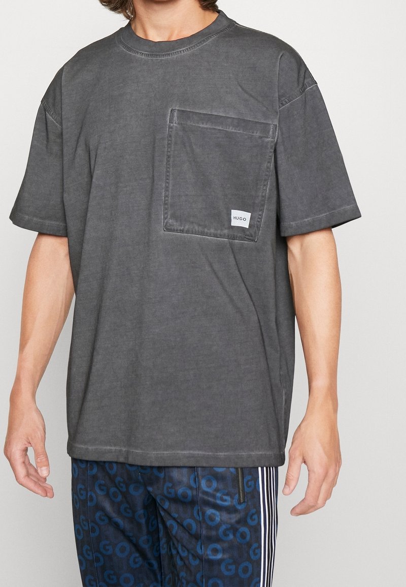 Homme portant un t-shirt noir délavé oversize avec une grande poche poitrine étiquetée « Hugo » et un pantalon bleu à motifs avec des rayures blanches.