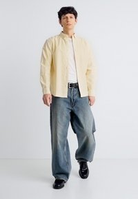 Jeune homme marchant en jean large bleu, chaussures noires, t-shirt blanc et chemise à boutons jaune clair sur fond blanc uni.