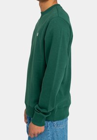 Uomo che indossa un semplice felpa verde scuro con polsini e orlo a coste, abbinata a jeans di denim azzurro, mostrato di lato.