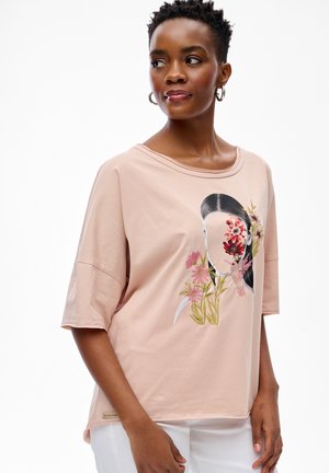 Kobieta o krótkich kręconych włosach, ubrana w jasnoróżową koszulę z kwiatowym i ptasim motywem oraz białe spodnie, stojąca na białym tle.