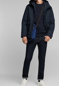 Donkerblauwe gewatteerde jas met capuchon, gecombineerd met een navy gebreide trui en donkere denim jeans. Persoon draagt een blauwe tas en witte sneakers.