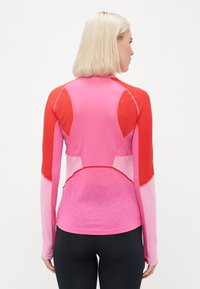 Kobieta z blond włosami, ubrana w długą różowo-czerwoną bluzę sportową i czarne legginsy, stoi tyłem na białym tle.
