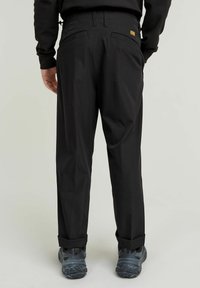 Svarta byxor med en avslappnad passform, med en knäppning på bakfickan och en diskret logotyp. Tyget verkar slätt och hållbart.