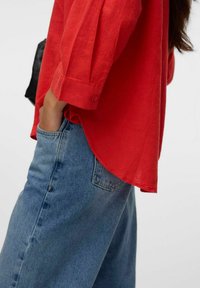 Blouse en lin rouge à coupe décontractée et manches trois-quarts, associée à un jean en denim bleu clair avec une poche avant et des coutures visibles.