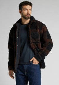 Brun och svart rutig fleecejacka med en strukturerad yta, knappframsida och bröstficka, bärs över en marinblå t-shirt och denimjeans.