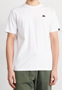 Hvid bomulds t-shirt med korte ærmer, med et lille flerfarvet logo på den øverste venstre bryst og et sort mærke på den højre ærme.
