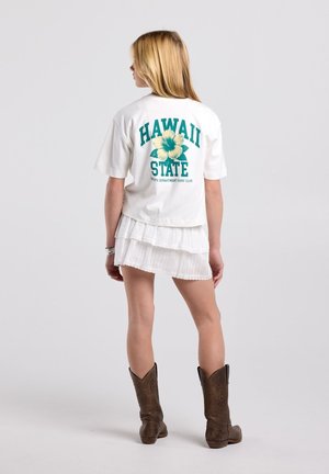 Meisje staat met haar rug naar de camera, draagt een wit T-shirt met opdruk "Hawaii State", een witte gelaagde rok en bruine cowboylaarzen op een effen achtergrond.