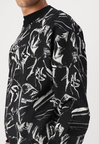 Black sweatshirt with white abstract patterns, featuring a ribbed neckline and a logo print on the lower side. Textured fabric finish.