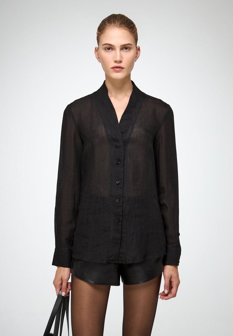 Camicia nera trasparente con colletto alla coreana, maniche lunghe e una sottile texture. Abbinata a pantaloni corti in pelle nera.