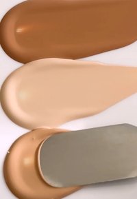 Tre prøver av flytende foundation: lys beige, middels beige og dyp varm brun, med en metallspatel som hviler på den middels nyansen.