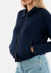 Felpa blu navy con zip e colletto, caratterizzata da un ricamo "BM" sul petto e tasche frontali. Tessuto morbido e design aderente.