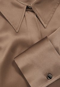 Chemise en satin marron avec col pointu et poignets avec un seul bouton noir brillant, mettant en valeur la texture lisse du tissu et des coutures soignées.