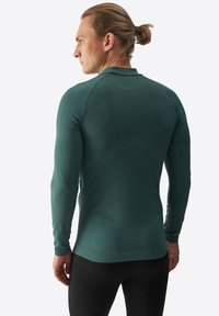 Camiseta deportiva de manga larga en verde oscuro, confeccionada en tejido texturizado, con un corte ajustado y costuras planas para mayor comodidad. Detalles con un sutil logotipo.