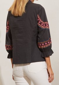 Blouse noire avec motifs brodés colorés sur les manches, dotée de poignets froncés et d'une coupe décontractée. Matière en coton léger texturé.