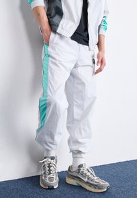 Bílé tepláky s tyrkysovými bočními akcenty, elastickými manžetami a lehkým materiálem. Nosené s šedými teniskami a námořnickým tričkem.