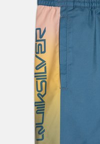 Modro-rumene kratke kopalne hlače z elastičnim pasom, stransko žepom in izrazito temno modro logotipom na strani.