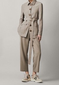 Beżowa kurtka z paskiem i dużymi guzikami, z dwoma przednimi kieszeniami, zestawiona z luźnymi, beżowymi spodniami i jasnymi trampkami, tworząc skoordynowaną stylizację.