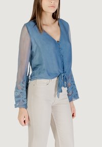 Blusa azul clara con escote en V, cierre de botones y detalle de lazo. Las mangas largas transparentes presentan un intrincado bordado azul y sutiles acentos de cuentas.
