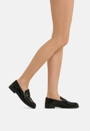 Gambe con mocassini neri lucidi con motivo coccodrillo e tacchi bassi, viste di profilo su uno sfondo chiaro e uniforme.