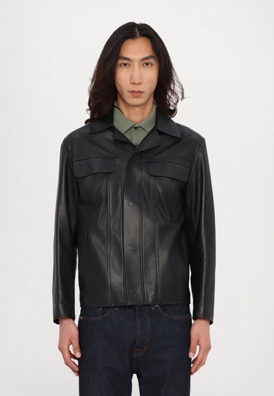 Giacca di pelle nera con bottone frontale, due tasche sul petto e cuciture strutturate. Indossata sopra una camicia verde con colletto.