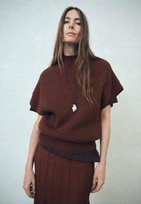 Femme aux longs cheveux bruns portant un pull en maille marron foncé à manches courtes et une jupe plissée assortie, avec un collier à pendentif.