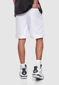 Shorts d'athlétisme en maille blanche avec poches latérales et coupe décontractée, associés à des baskets noires et blanches arborant un logo bien visible.