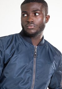 Veste bomber bleu marine avec une texture lisse, fermeture éclair, poignets et col côtelés. Présente un marquage discret dans le tissu.