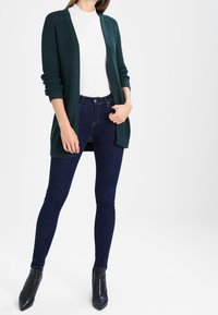 Dunkelgrüne Strickjacke mit offenem Vorderteil, strukturiertem Muster, kombiniert mit enganliegenden dunklen Jeans und schwarzen Stiefeletten.