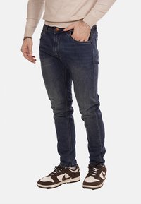 Jeans in denim blu scuro con una vestibilità slim, dotati di cinque tasche e cuciture arancioni a contrasto. Abbinati a sneaker marroni e bianche.