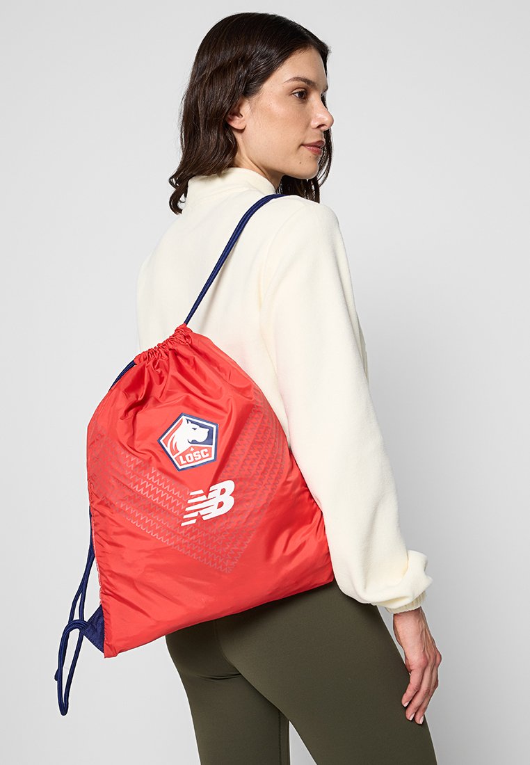 Czerwony plecak na sznurku wykonany z nylonu z diagonalnymi wzorami. Posiada naszywkę z logo i branding "New Balance" w białym kolorze. Granatowe sznurki.