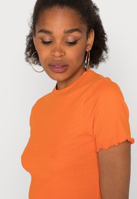T-shirt laranja, ajustada, feita de tecido suave. Apresenta mangas curtas com frufrus e um decote redondo clássico. Cor clara e vibrante.