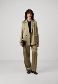 Oliivinvihreä puku, jossa kaksirivinen bleiseri ja cargo-housut. Sileä pinta, monikäyttöinen design, yhdistetty mustaan toppiin ja clutch-laukkuun.