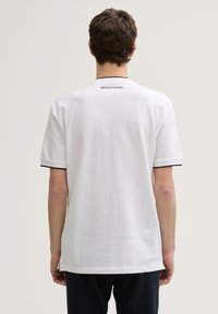 Tricou alb cu mânecă scurtă, cu guler rotund și detalii negre pe mansete. Are branding pe partea superioară a spatelui. Material moale, croi lejer.