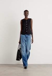 Svart ermeløs topp med knapper, paret med løse blå jeans. Modellen bærer en svart quiltet håndveske og har på seg spisse svarte sko.