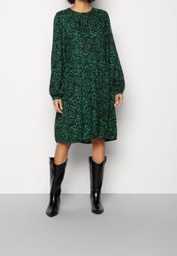 Vestido verde con estampado y diseño abstracto negro, de mangas largas y cuello redondo. Combinado con botas negras de charol hasta la rodilla.