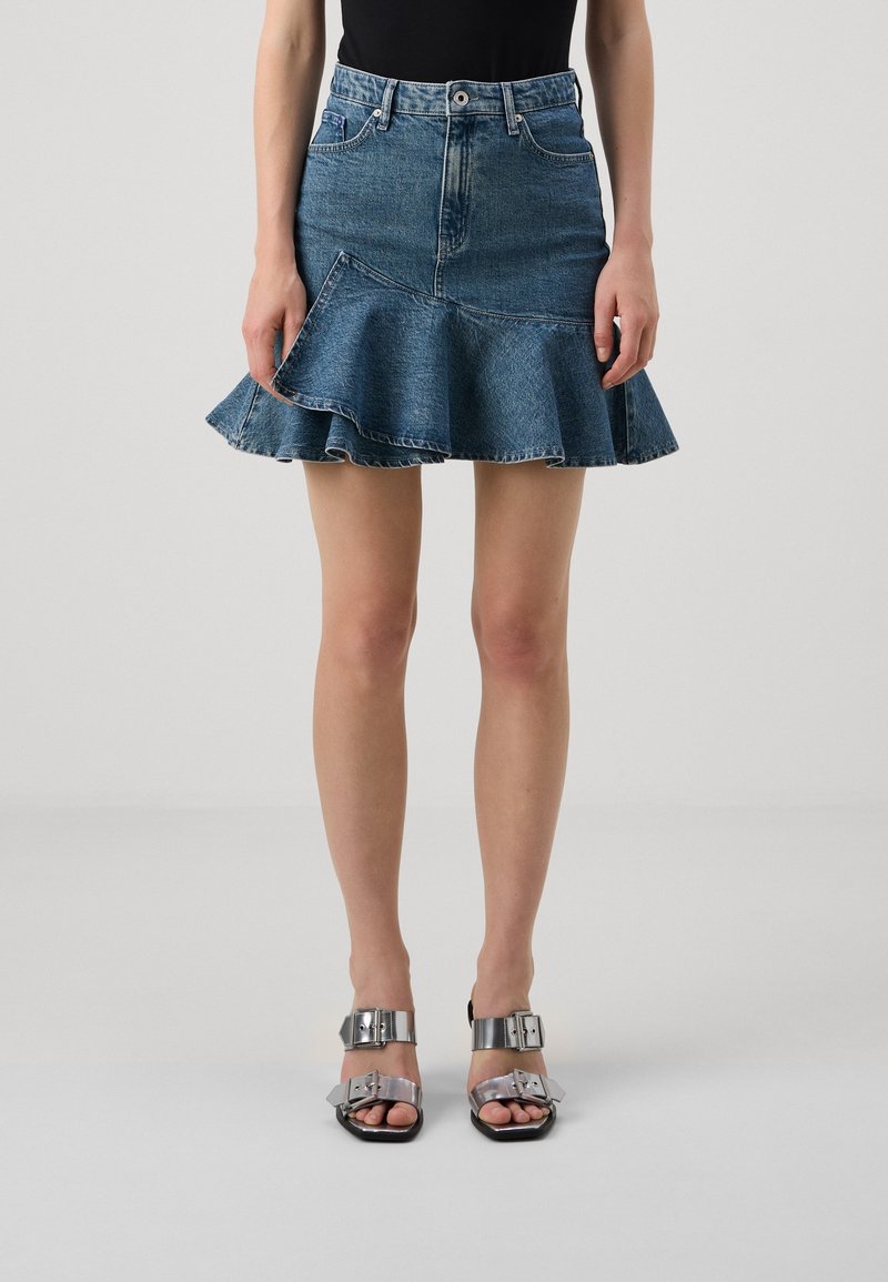 Vrouw in een blauwe denim mini-jurk met ruches, gecombineerd met een zwarte top en sandalen met een zilveren gesp, staand tegen een effen achtergrond.