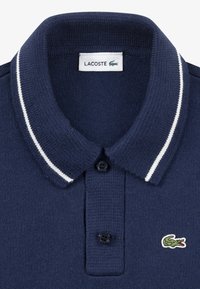 Ναυτικό μπλε πόλο μπλουζάκι από πλεκτό ύφασμα, με πλάκα δύο κουμπιών, γιακά με λευκές ρίγες και πράσινο λογότυπο κροκόδειλου.