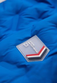 Blauwe gewatteerde stof met een doorzichtige patch met het label "ombre performance" en een rood-wit-zwart chevronpatroon.