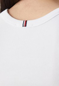 Tessuto bianco a coste con scollo ampio, caratterizzato da un piccolo dettaglio di strisce rosse e blu navy sul retro del collo. Texture liscia e aderente.