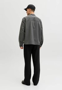 Chaqueta camisa gris de corte relajado y mangas largas, combinada con pantalones negros con rayas finas y zapatos negros, vista desde atrás.