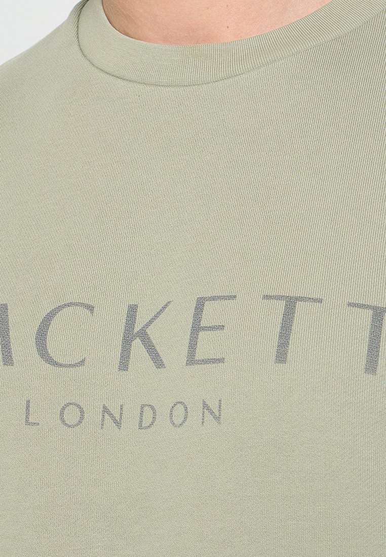 Gros plan sur une chemise en tissu côtelé vert clair avec un texte gris indiquant "Hackett London" sur la poitrine.