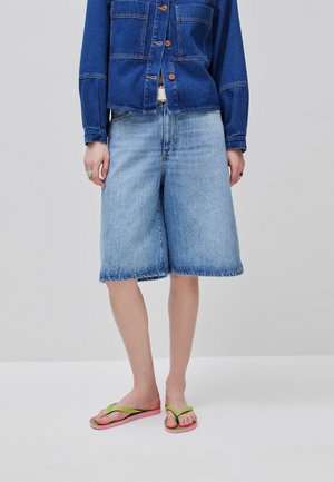 Personne portant une veste en jean bleu foncé, un short large en jean bleu clair, et des tongs vertes et roses, debout sur un fond uni.